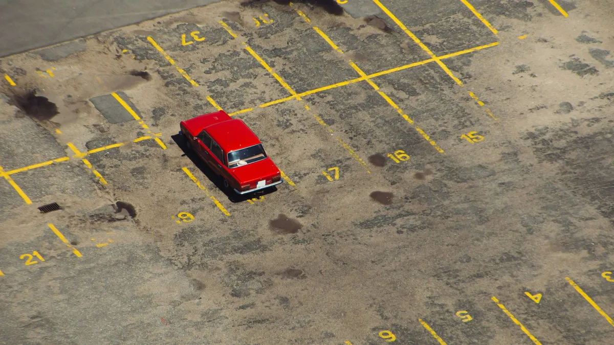 Carro vermelho antigo estacionado sozinho num estacionamento vazio, visto do alto, com vagas numeradas pintadas de amarelo