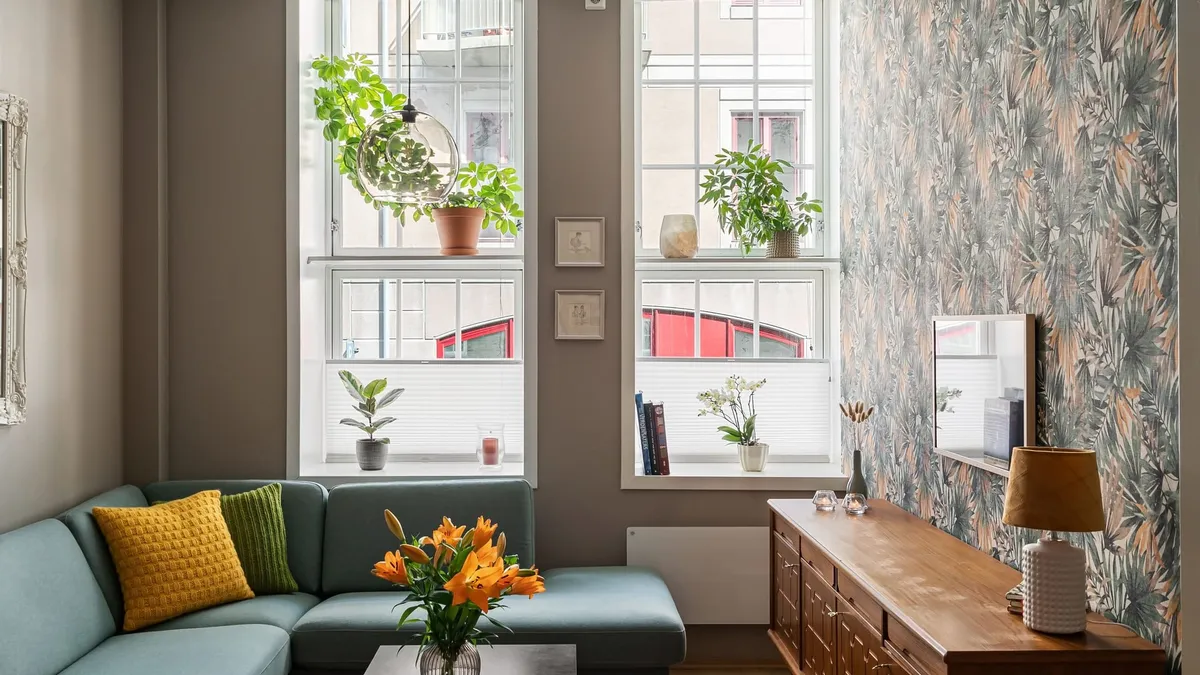 Sala de apartamento com sofá verde de canto, almofadas amarela e verde, flores laranja na mesa de centro, duas janelas altas com plantas e parede com papel de folhagens